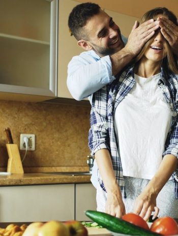 Young man covering girlfriends eyes with hands and surprising her in the kitchen at home. Loving couple cooking breakfast in the morning
