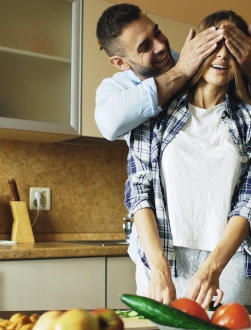 Young man covering girlfriends eyes with hands and surprising her in the kitchen at home. Loving couple cooking breakfast in the morning