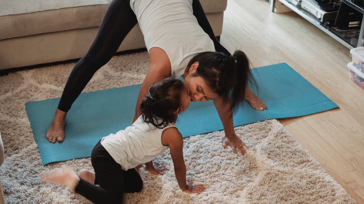 Positive fit Asian female in activewear performing Downward Facing Dog pose and kissing adorable daughter while practicing yoga in living room